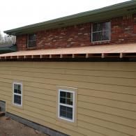 A tan-sided room addition under construction with exposed rafters, attached to a brick home with two new windows.