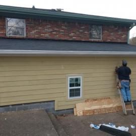 A person on a ladder installs yellow siding on the exterior of a brick and yellow house.
