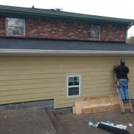 A person on a ladder works on the beige siding of a house with a brick upper story and a new dark shingle roof.