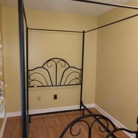 A black metal four-poster bed frame with an arched, decorative scroll-patterned headboard, placed in a room with tan walls.