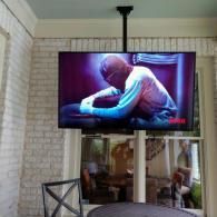 A flat-screen TV mounted from the ceiling on a patio, displaying a person leaning forward against a purple background.