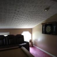 A brown leather sofa sits in a room with a sloped, textured ceiling, pink carpeting, and a wall-mounted dartboard.