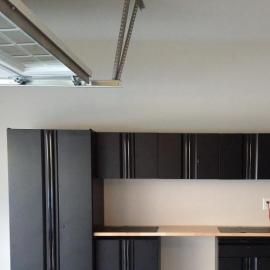 A row of dark cabinets with a light-colored countertop in a garage setting, beneath a metal garage door track.