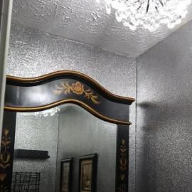 A decorative black mirror with gold floral accents hangs on a wall covered in shimmering silver textured wallpaper.
