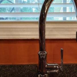 A chrome kitchen faucet stands against an orange backsplash in front of a window with closed white blinds.