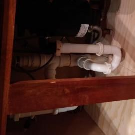 White PVC plumbing pipes and a black garbage disposal unit installed under a wooden kitchen cabinet.