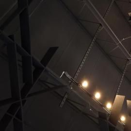 Low-angle view of industrial metal ceiling rafters illuminated by a row of warm, round track lights against a dark ceiling.
