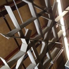 A low-angle view of a grey, geometric lattice structure hanging from a ceiling, composed of interlocking angular panels.