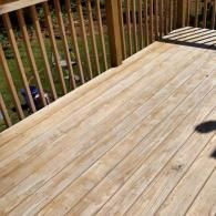 A high-angle view of a wooden deck with light brown, parallel floorboards and vertical railing posts.