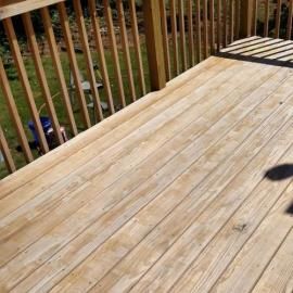 A top-down view of a light-colored wooden deck with vertical railing, seen from an elevated angle on a sunny day.