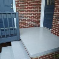 A set of two gray concrete steps leading to a gray porch with a blue railing and two blue doors against a brick wall.