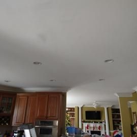 A kitchen with dark wood cabinets and recessed ceiling lights, looking toward a living room with a fireplace.