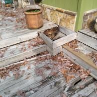 Worn wooden deck steps with a missing plank, exposing the hollow interior, surrounded by fallen autumn leaves.