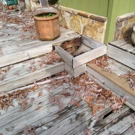 A weathered wooden deck showing significant rot, missing boards, and an open, damaged section near the stairs.