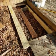 An open section of a wooden deck reveals joists and ground cover, with some construction materials nearby.