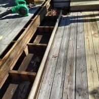 A partially removed wooden deck showing the support joists, with a green leaf blower on the left.