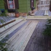 A green house with a multi-level wooden deck featuring a mix of new, light-colored boards and older, weathered gray boards.