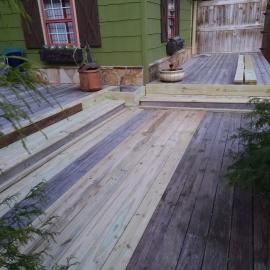A wooden deck transitions from weathered, grey boards to new, lighter-colored planks, leading toward a green house exterior.