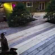 A wooden deck showing a section of new, lighter-colored replacement boards contrasted with older, weathered gray planks.