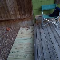 A wooden ramp leads from a dirt yard up to a weathered wooden deck with a green folding chair near a green wall.