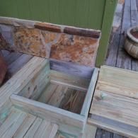A wooden planter box sits on a deck, positioned against a house wall with a stone veneer foundation.