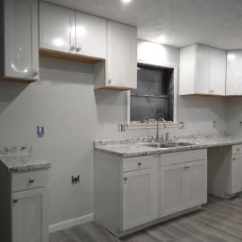 A bright, modern kitchen features white cabinets, light speckled countertops, a sink, and grey wood-look flooring.