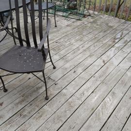 A weathered wooden deck with a metal chair and parts of a table visible, overlooking a patch of dirt and sparse greenery.