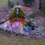 A backyard stone pond with a small waterfall, illuminated by vibrant, multicolored lights reflecting on the water.