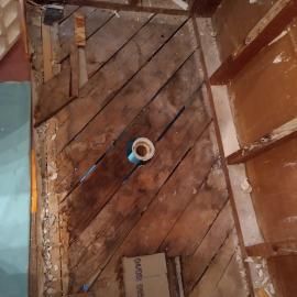 A partially demolished bathroom floor showing exposed wood planks and a central drain pipe.