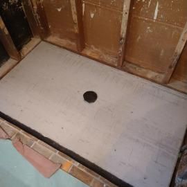 A newly installed concrete shower floor pan with a central drain hole, set against wooden wall studs in a room under remodel.