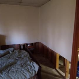 A room with a bed and unfinished walls where the lower portion of the drywall has been removed, exposing the framing.