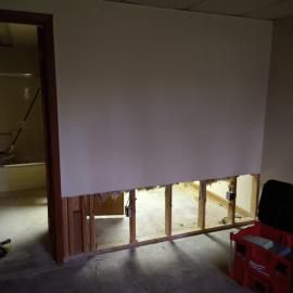 A room with a partially demolished wall exposing wooden studs near the floor, with a doorway leading to a bathroom.