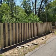 A wooden fence with alternating light and dark pickets stands along a paved road next to lush green trees.