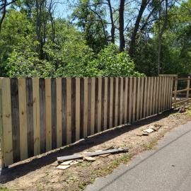 A wooden privacy fence made of alternating light and dark panels stands beside a paved road with trees in the background.