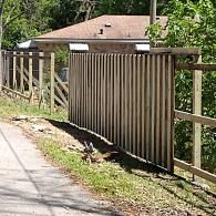 A newly constructed wooden fence with vertical pickets runs along a gravel path in front of a brick house.