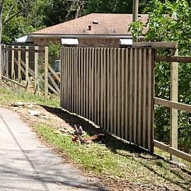 A wooden fence under construction with vertical slats along a dirt path near a house.