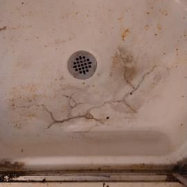 A dirty, off-white shower floor with significant cracks radiating from near the drain and accumulated grime and rust.