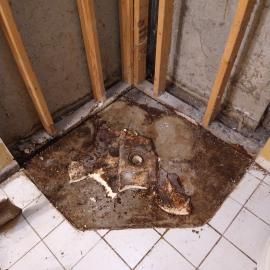 A corner of a room with exposed wall studs and a damaged, stained subfloor surrounding a central pipe hole.
