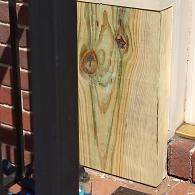 A rectangular piece of light-toned, wood-grain planking sits upright on a brick patio next to a black metal railing.