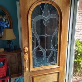 A wooden front door with a tall, arched decorative glass panel featuring a swirled, etched design and a gold handle.