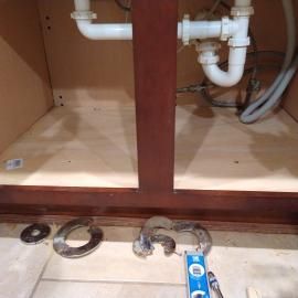Under-sink cabinet with white plumbing pipes, tools, and metal hardware lying on the floor in front of the cabinet.