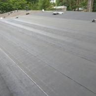 A flat commercial roof with grey membrane surfacing, showing seams between rolls and distant tree lines.