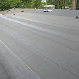 A view of a flat residential roof covered in grey, rolled membrane material, with trees in the background.