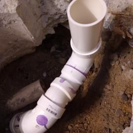 A white PVC plumbing assembly with purple primer stains, positioned in a dirt trench beside concrete.