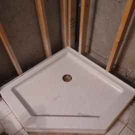 A white, pentagonal shower base installed in a corner with unfinished wooden wall studs in the background.