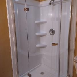 A white shower stall with glass doors, built-in corner shelves, and a metal shower head and handle.