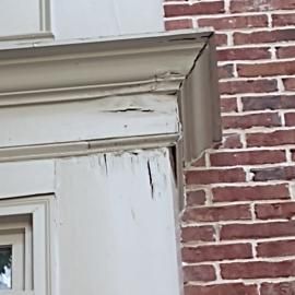 Peeling white paint and wood rot on the decorative exterior trim and corner post of a brick building.