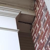 A white pillar top meets a brick wall, showing a noticeable gap where the wooden trim has separated from the brick.