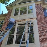 A person on a ladder repairs the wood trim between the upper and lower windows on a two-story brick house exterior.