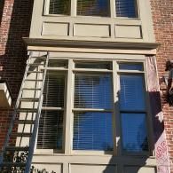 A tall ladder leans against a brick house exterior, beside a multi-pane bay window structure undergoing repair.